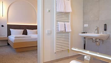 A hotel room with a bed, a bedside table, a wall-mounted heater, a towel rack, and a bathroom with a sink and mirror.