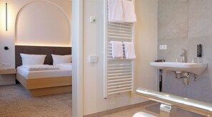 Una camera d'albergo con un letto, un comodino, un termosifone a parete, un portascopevoli e un bagno con lavandino e specchio.
