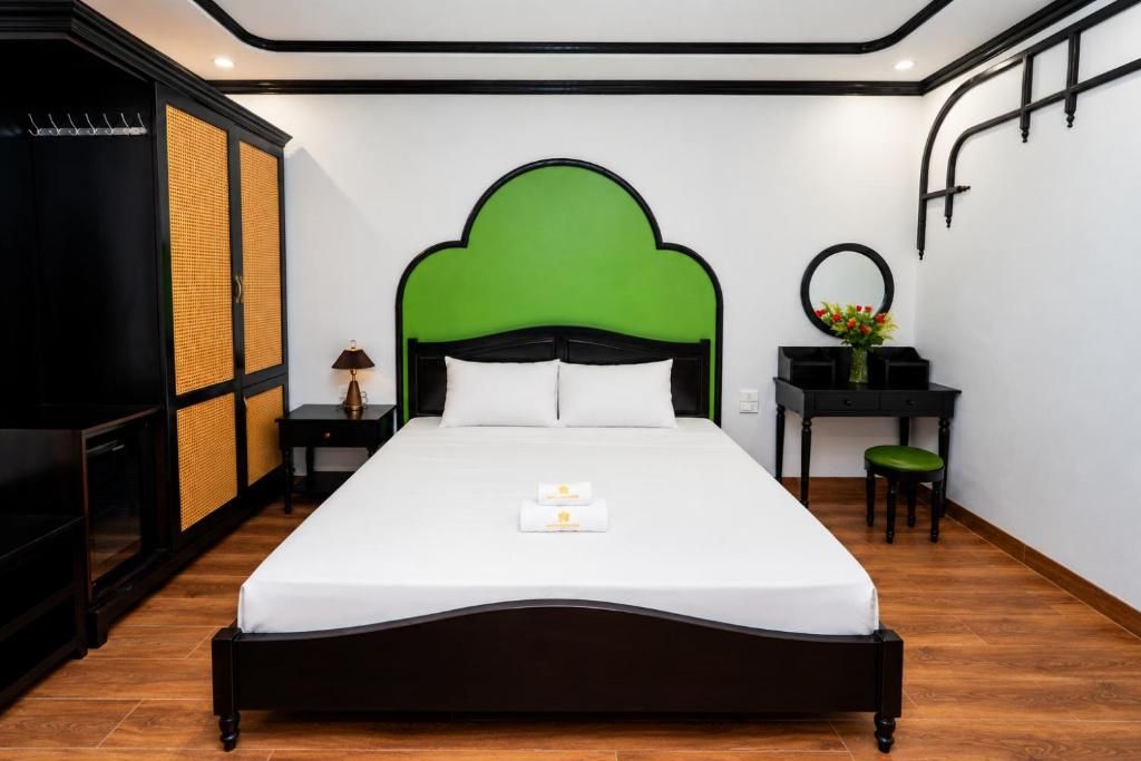 Una cama bien hecha con cabecera verde, una mesita de noche con lámpara y un pequeño escritorio con espejo y flores.