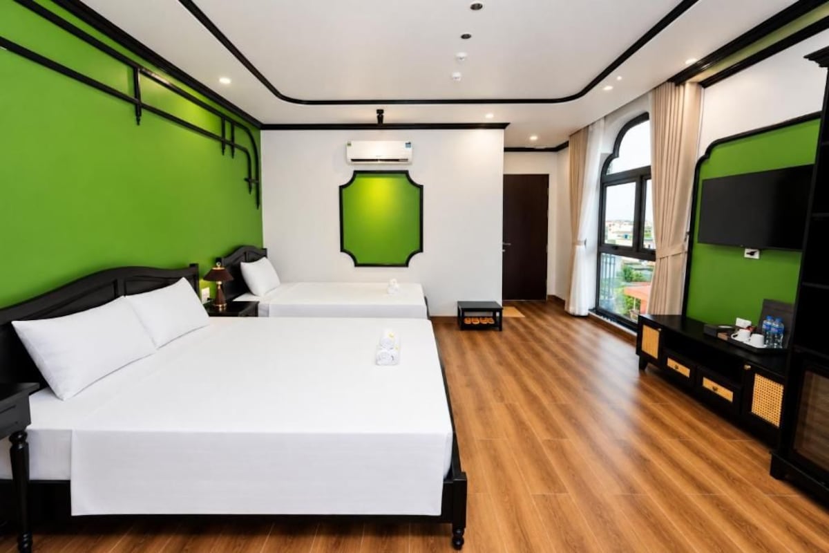 Habitación de hotel con dos camas, una pared de acento verde, un ventanal grande y un televisor sobre un soporte negro.
