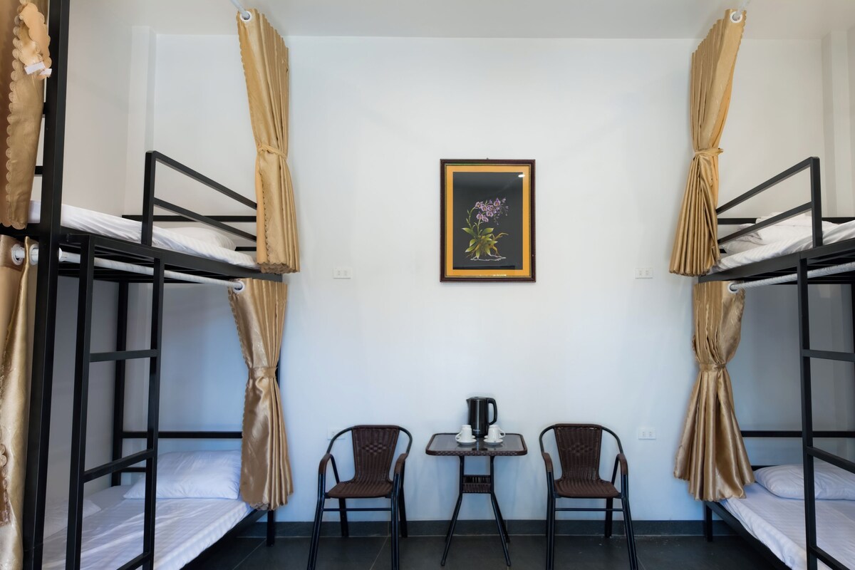 A dormitory room with bunk beds, a framed floral artwork on the wall, a small table with two cups, and two chairs.