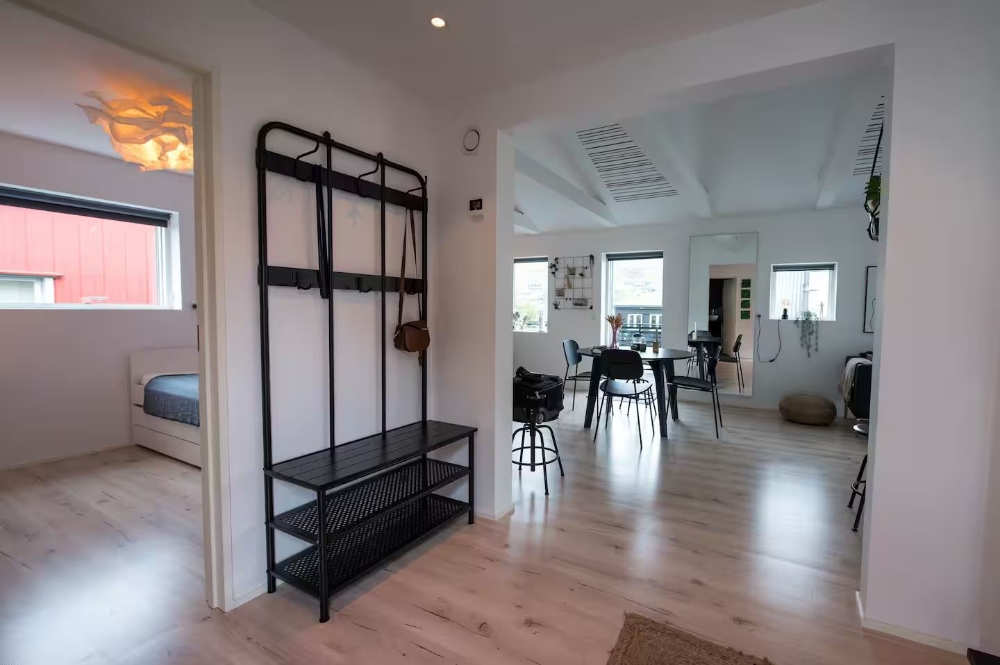 A spacious room with a bed, a black metal shelf unit, a dining area with chairs and a table, and a view through an open doorway.