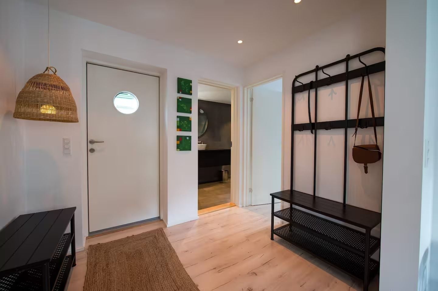 A hallway with a door, a hanging light fixture, a black metal shelf with a bag, and a wooden floor.