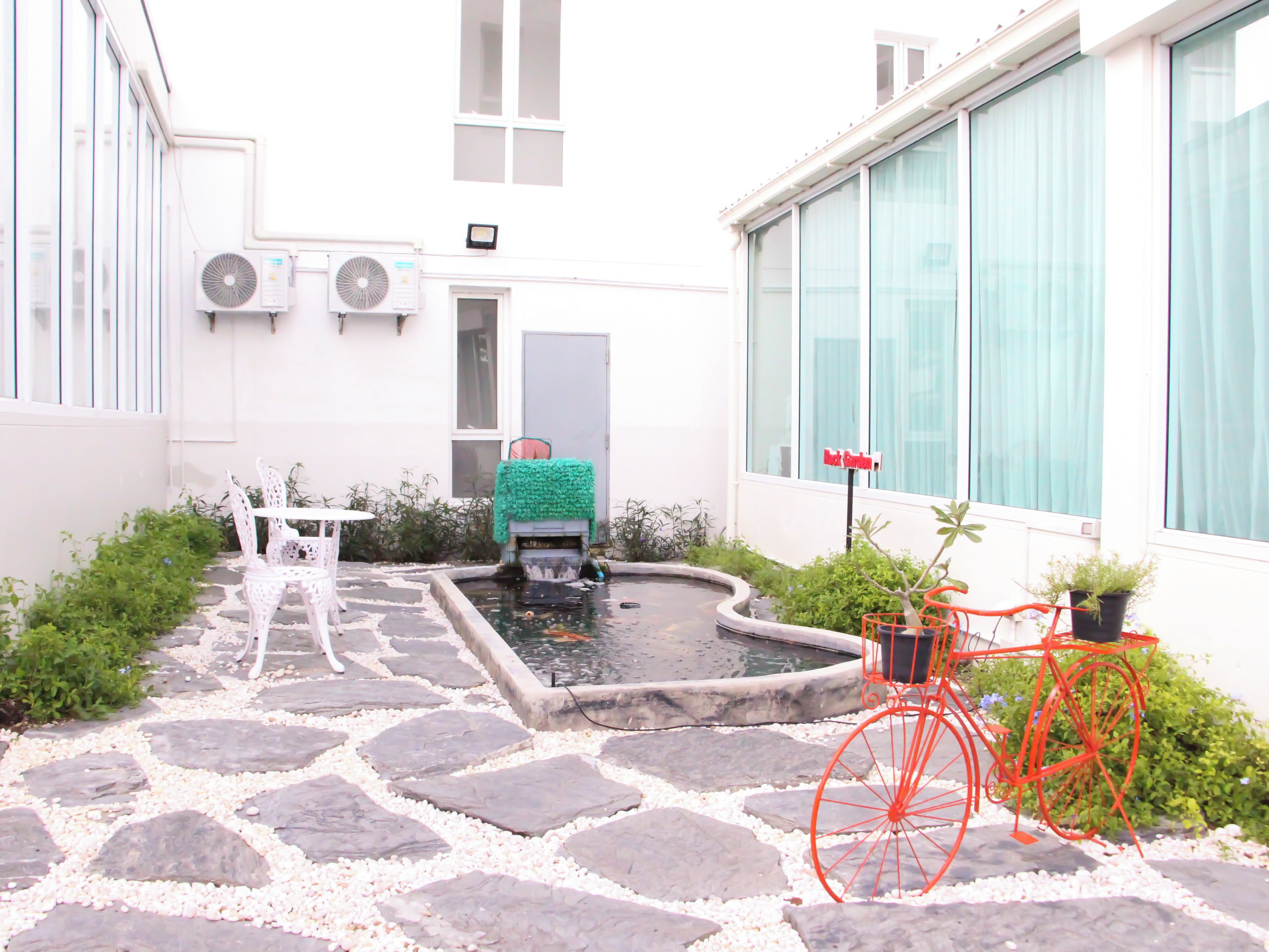 A courtyard with a stone pathway, a small pond, a red bicycle, and a white building with windows and air conditioning units.