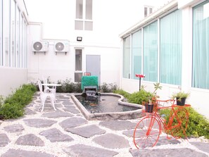 Een binnenplaats met een pad van stenen, een klein vijvertje, een rode fiets en een wit gebouw met ramen en airconditioners.