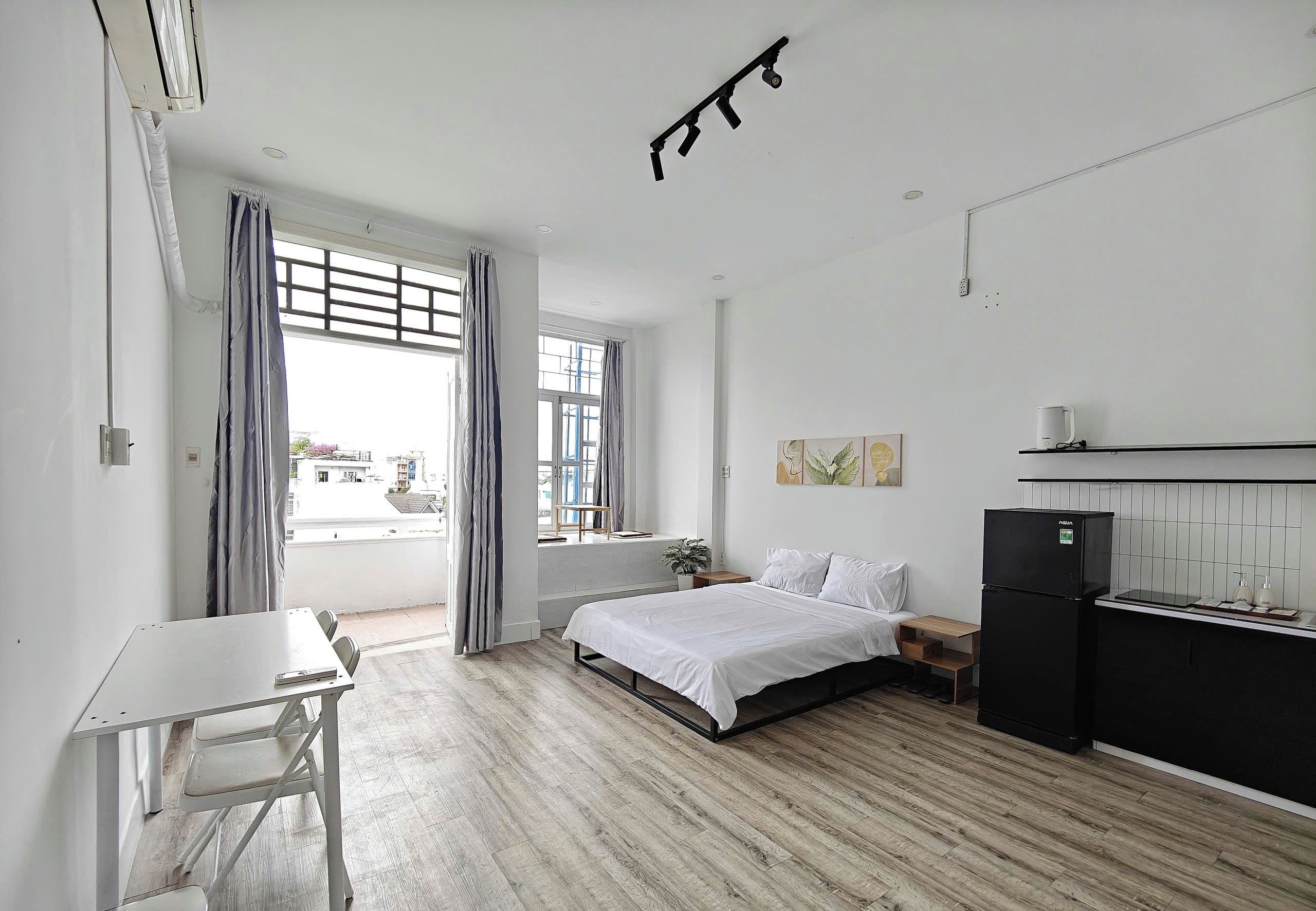 Un dormitorio minimalista con cama, escritorio, refrigerador y ventana con cortinas.