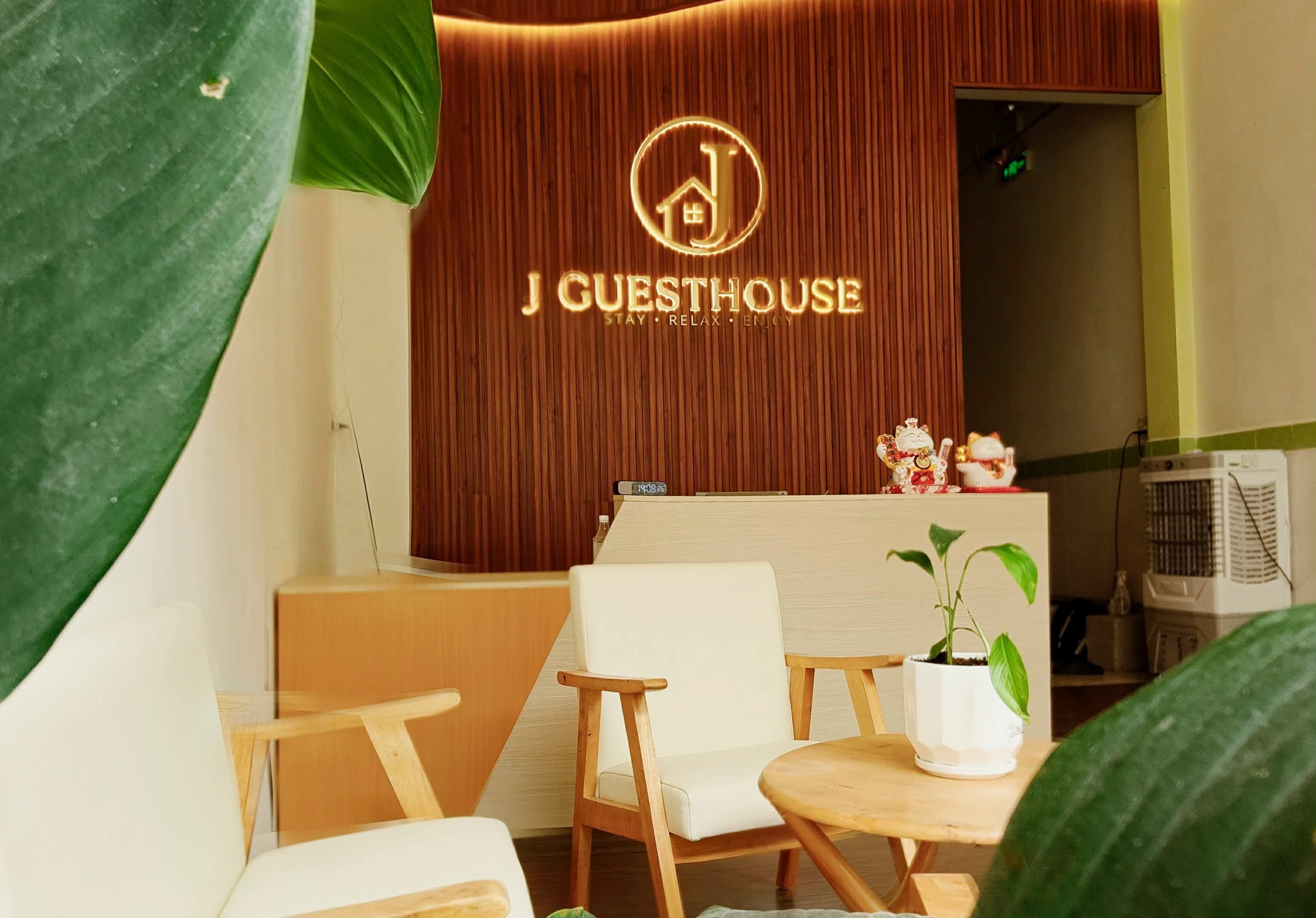 Una recepción con un panel de madera de fondo y el logo de 'J GUESTHOUSE'. Hay dos sillas y una mesita con una planta.