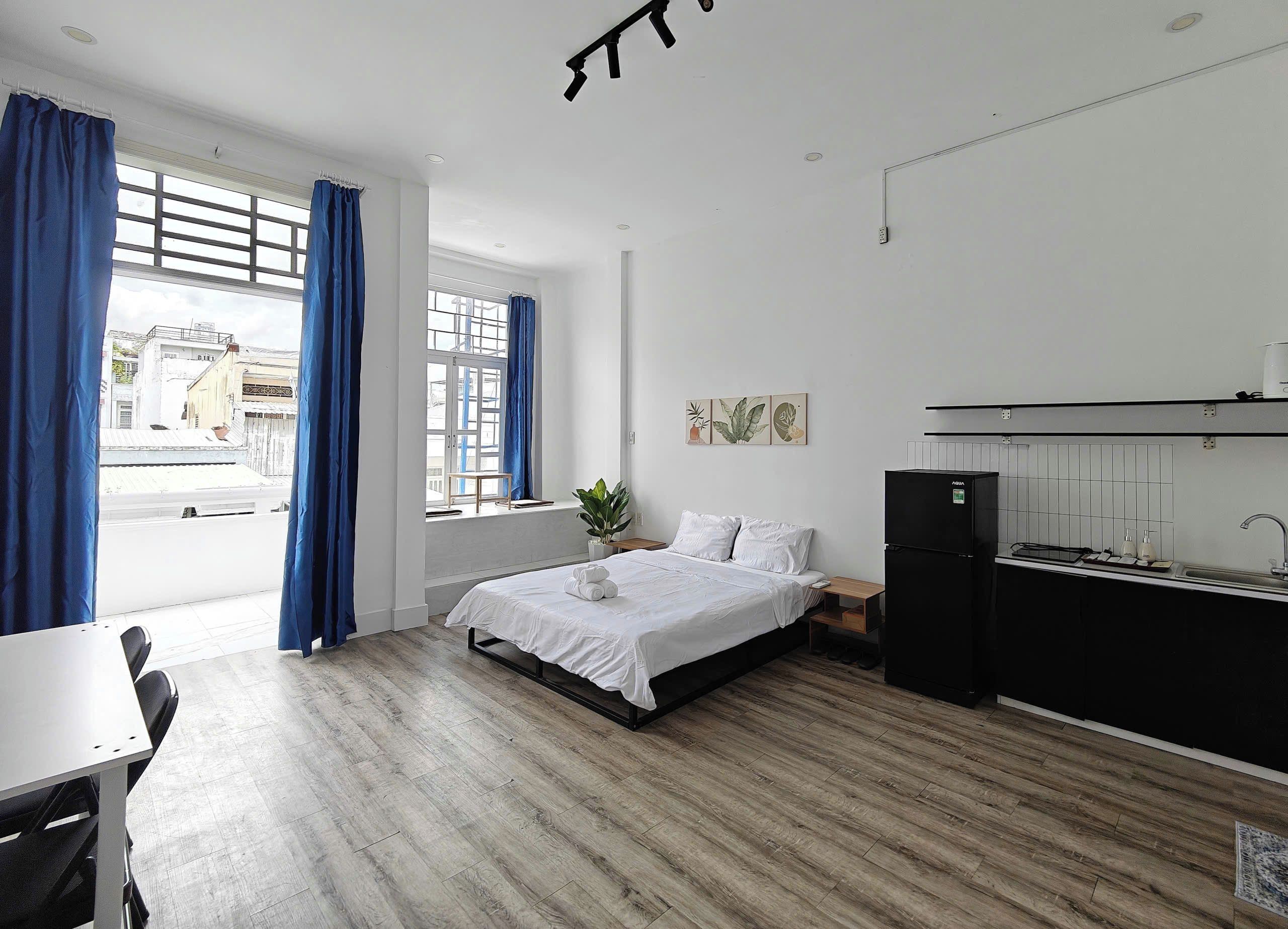 Un dormitorio con cama, escritorio, ventana con cortinas, planta y una kitchenette.