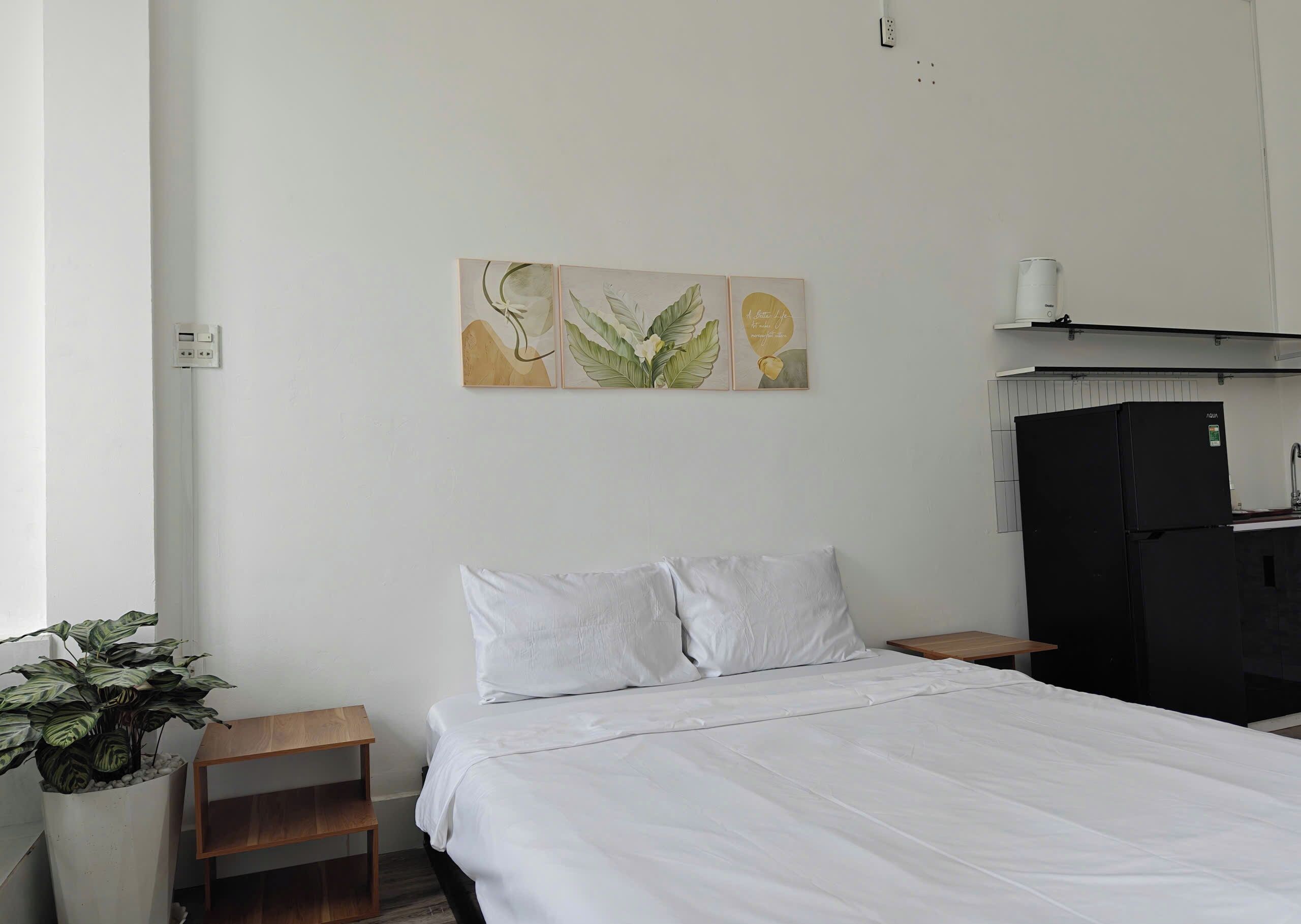 Un dormitorio con cama, ropa de cama blanca, una mesita de noche pequeña, una planta en maceta y una obra de arte colgada en la pared.