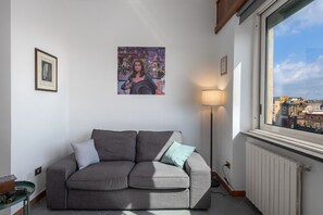 A living room with a grey sofa, a framed picture on the wall, a floor lamp, and a window overlooking a cityscape. - Toledo Skyview: Panoramic Penthouse in the Heart of Naples (Napoli)