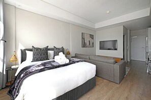 A modern bedroom with a large bed, grey sofa, and a flat-screen TV mounted on the wall. - Sleek Studio with City Skyline Views in Cape Town perfect for your getaway (Cape Town)