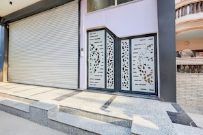 A closed metal shutter door with a geometric pattern, a set of stone steps leading up to a building entrance, and a balcony with a railing. - Collection O Sadar Bazar Gurgaon (Gurugram)