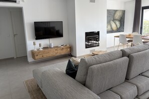 Ein modernes Wohnzimmer mit einem großen Flachbildfernseher, einem Holzkonsolentisch, einer Sitzecke mit grauem Sofa und einem Essbereich mit Tisch und Stühlen.