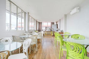 Uma área de refeições com mesas, cadeiras, janelas amplas e um aparelho de ar condicionado.