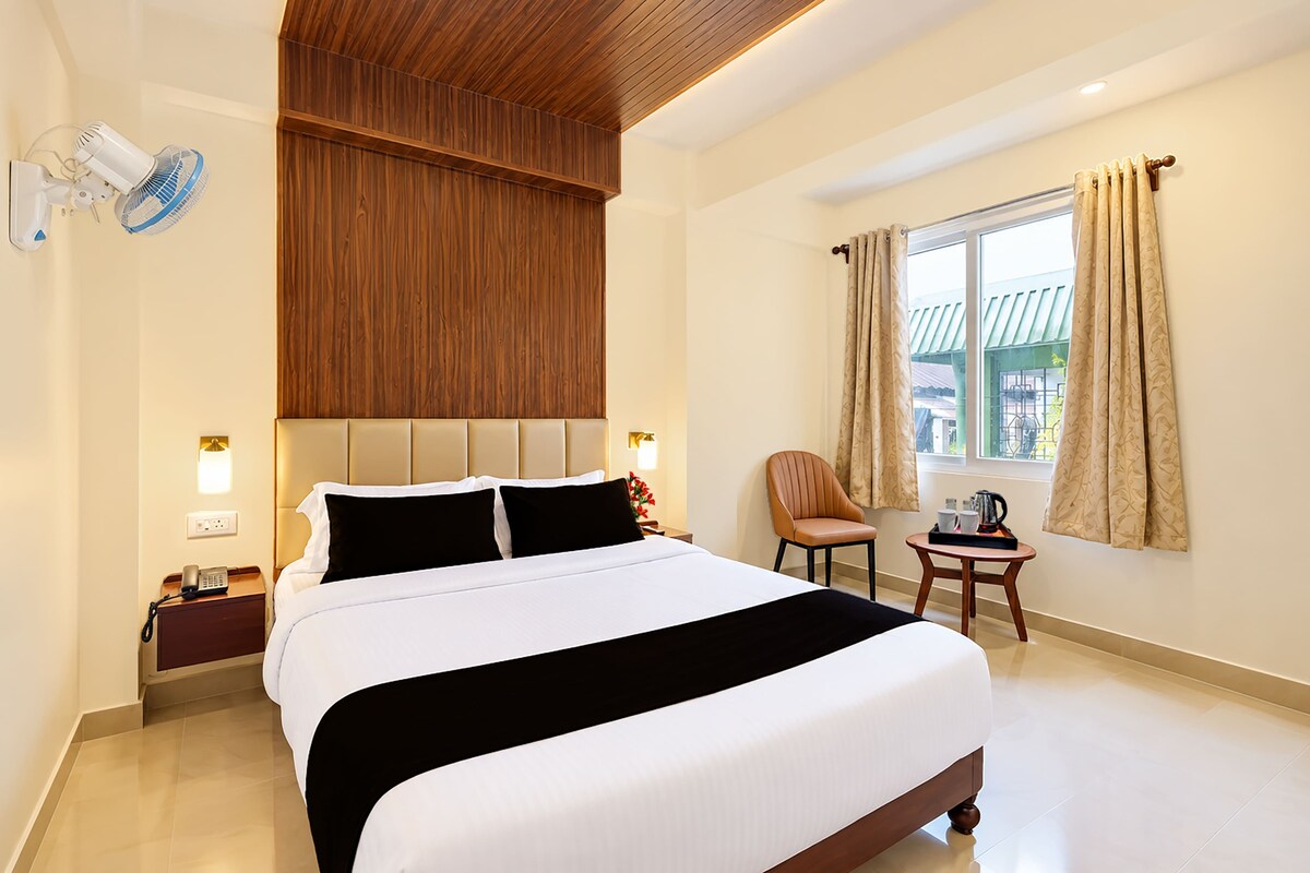 A hotel room with a large bed, a wooden headboard, a small table with a chair, a window with curtains, and a wall-mounted fan.