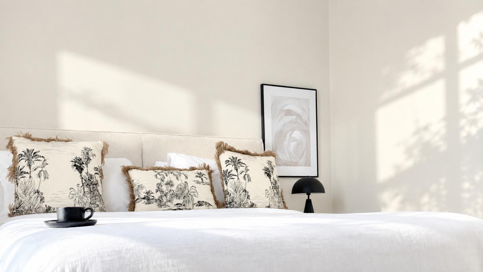 Una cama con una colcha blanca, tres almohadas con estampado tropical, una taza de café negro, una lámpara negra y un cuadro en la pared.
