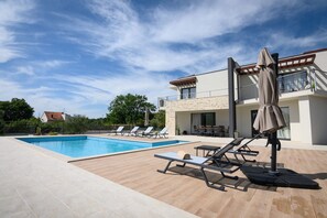Una piscina con sillas de descanso y sombrillas, un área de estar al aire libre techada, y un edificio de fachada de piedra.