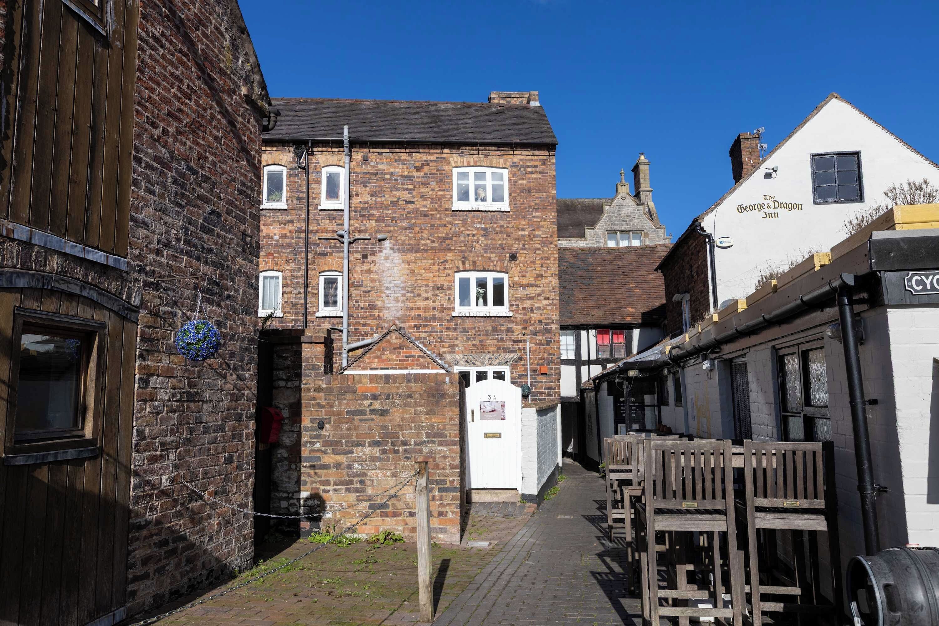 Eine schmale Straße mit traditionellen Backsteingebäuden, ein weißes Gebäude mit der Aufschrift 'The George & Dragon Inn' und ein Holzzaun auf der rechten Seite.