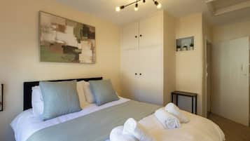 A bedroom with a bed, pillows, a blanket, a painting, a wardrobe, and a small table.