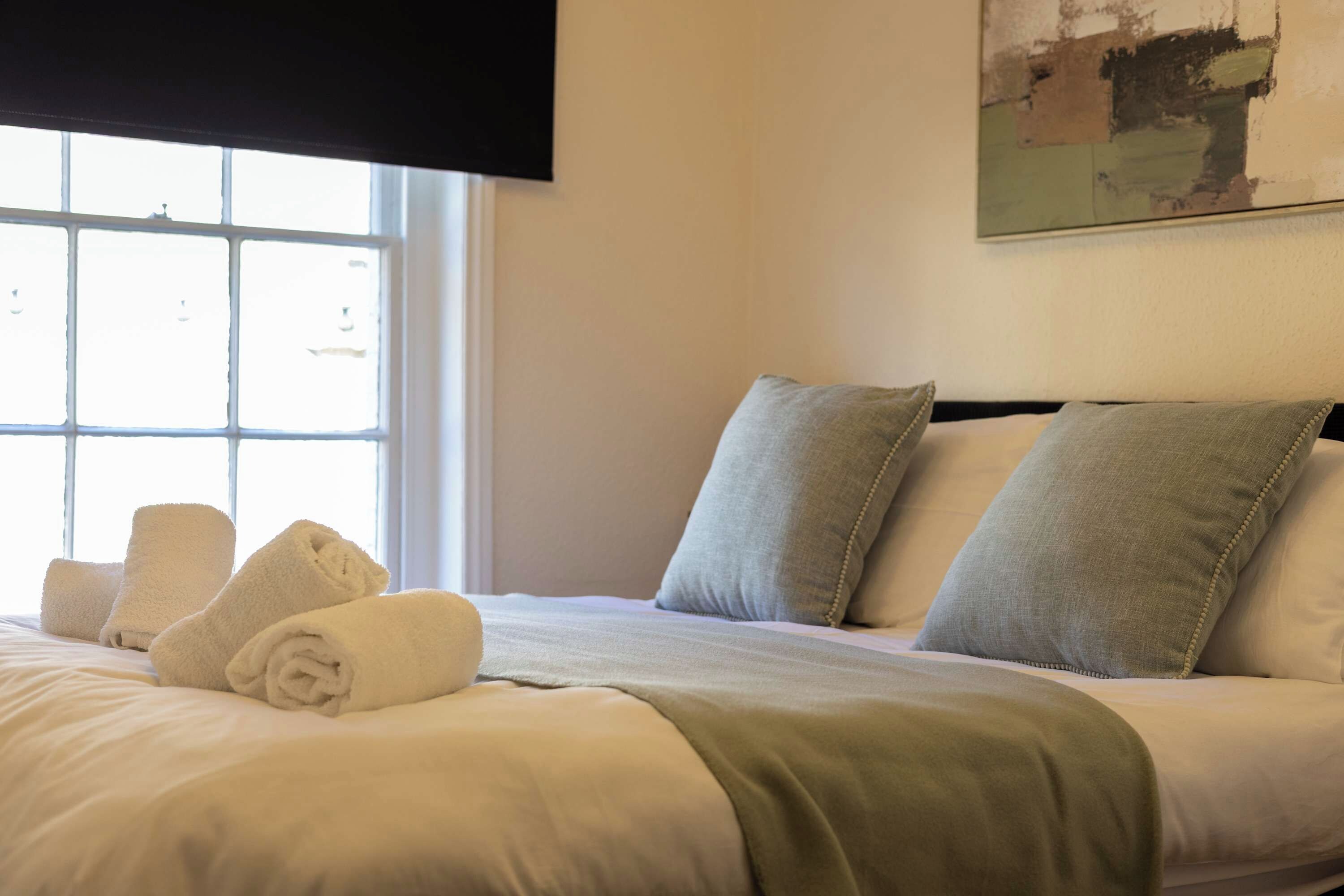 Ein ordentlich bezogenes Bett mit Kissen und aufgerollten Handtüchern, ein Fenster mit Jalousien und ein Bild an der Wand.