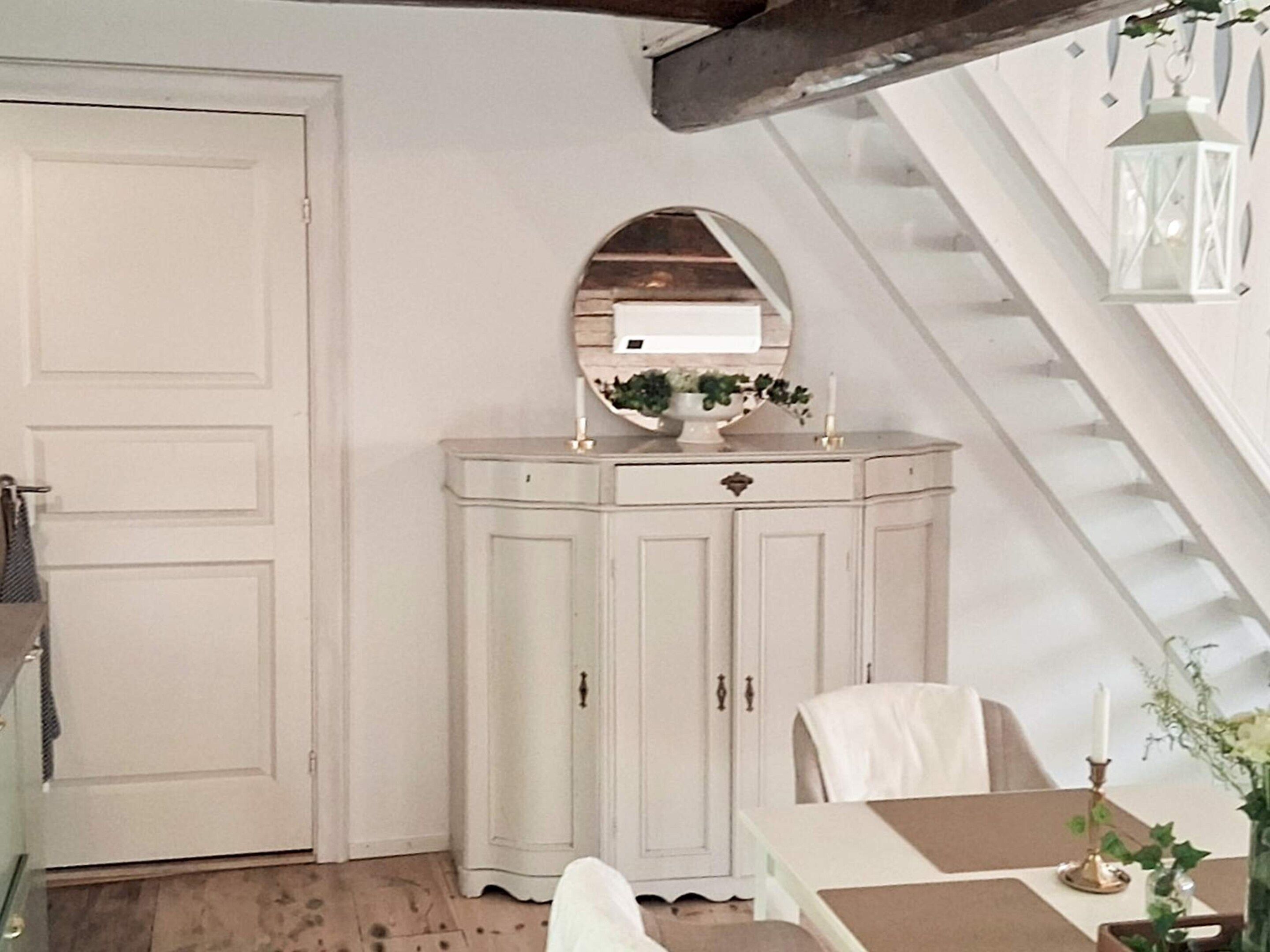 A room with a white door, a white cabinet with a mirror, a table with a vase, and a staircase.