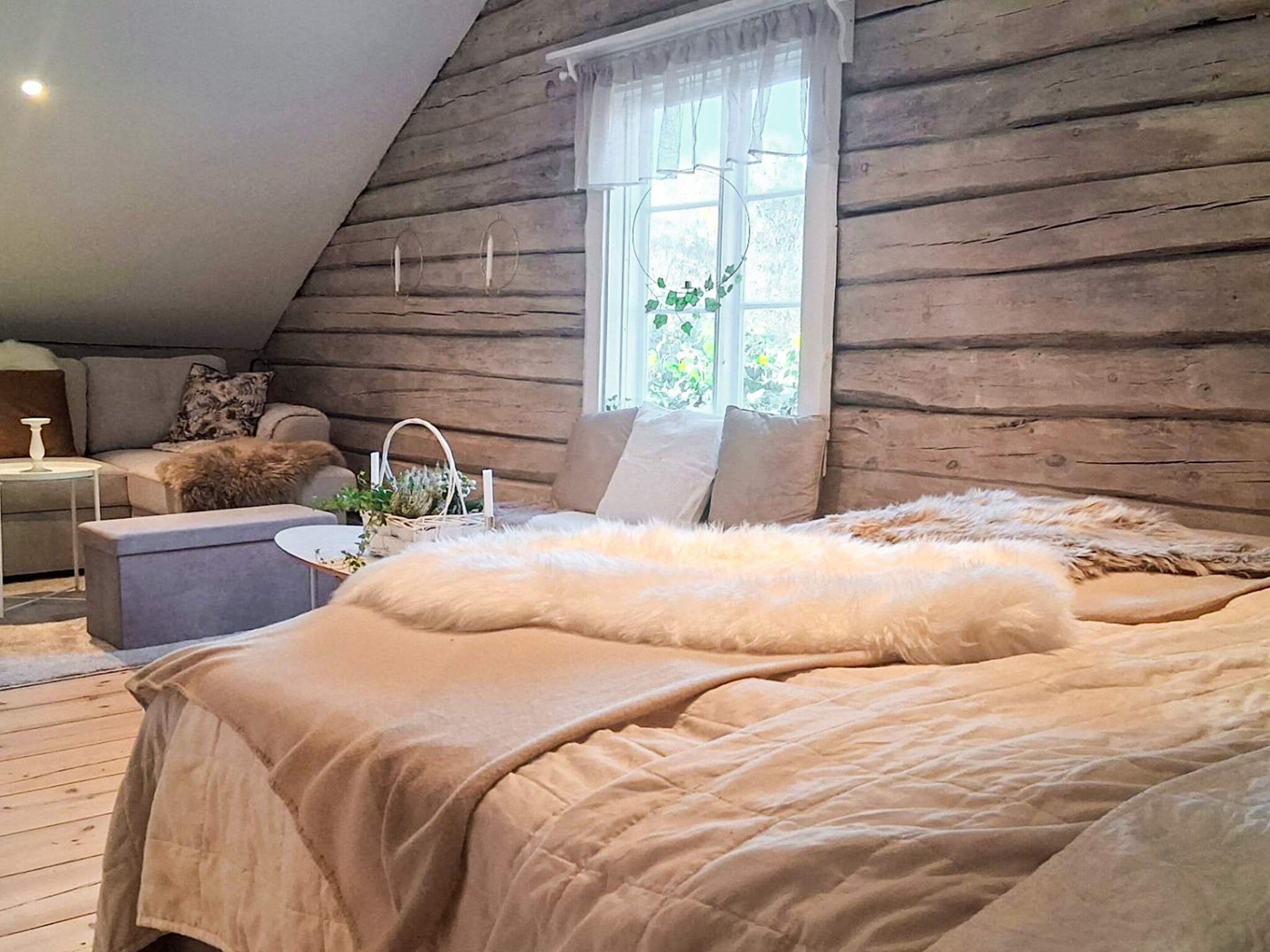 Ein gemütliches Schlafzimmer mit schrägen Wänden, einem großen Bett mit flauschiger Decke, einem Nachttisch mit Lampe und einem Fenster mit Vorhängen.