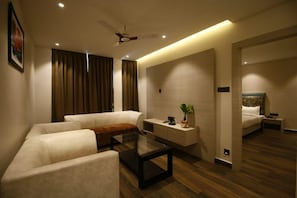 A modern hotel room with a sofa, a glass coffee table, a bedside table with a lamp, a bed with a headboard, and a ceiling fan. - The Pearl (Adityapur)