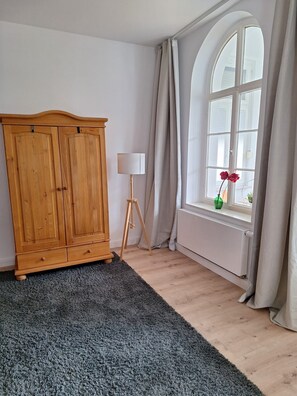 A room with a wooden wardrobe, a floor lamp, a window with curtains, and a rug.