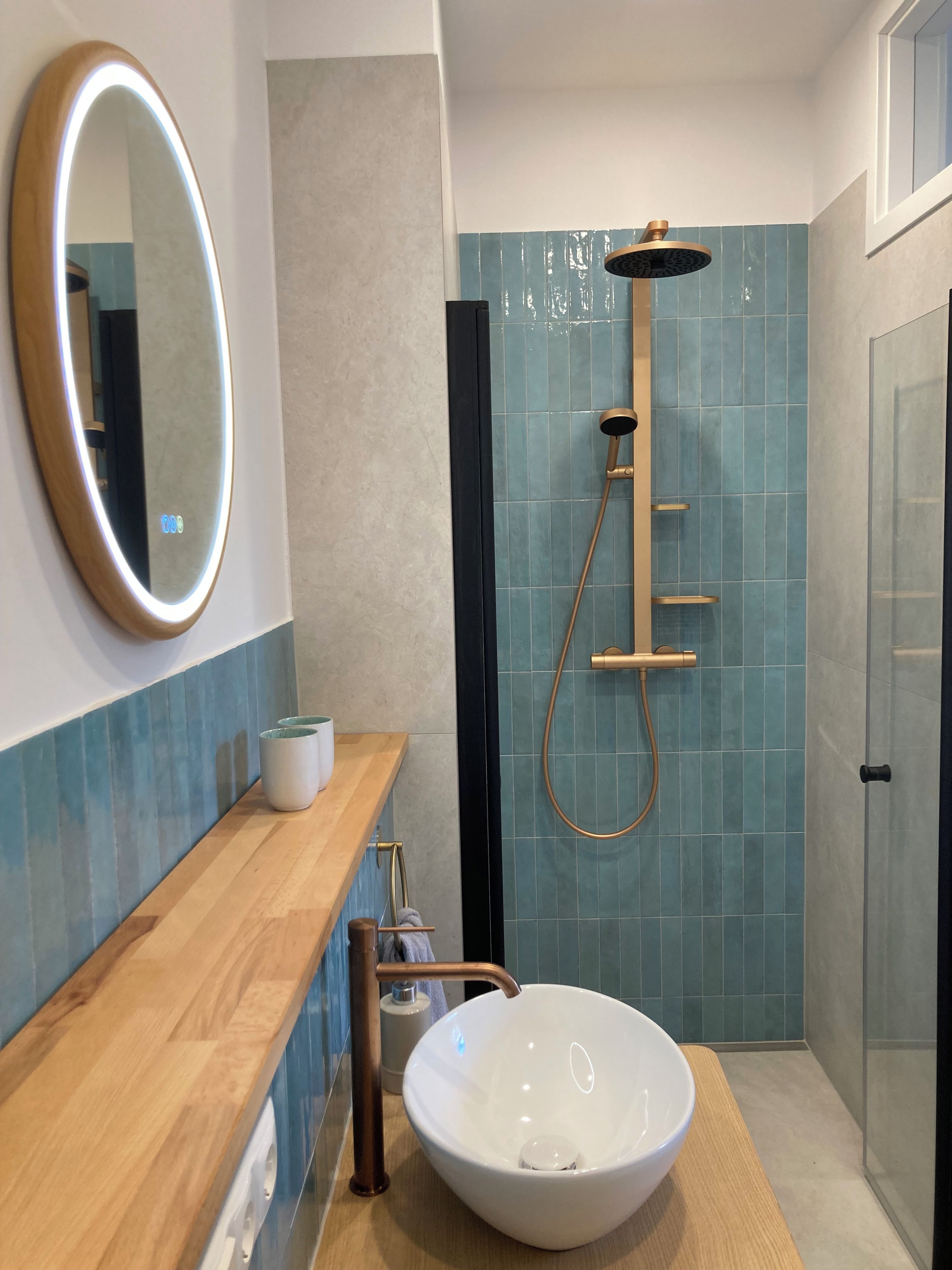 Ein Badezimmer mit einem Holzkosmetikschrank, einem großen, ovalen Spiegel, einem weißen Waschtischbecken und einer Dusche mit goldfarbener Armatur.