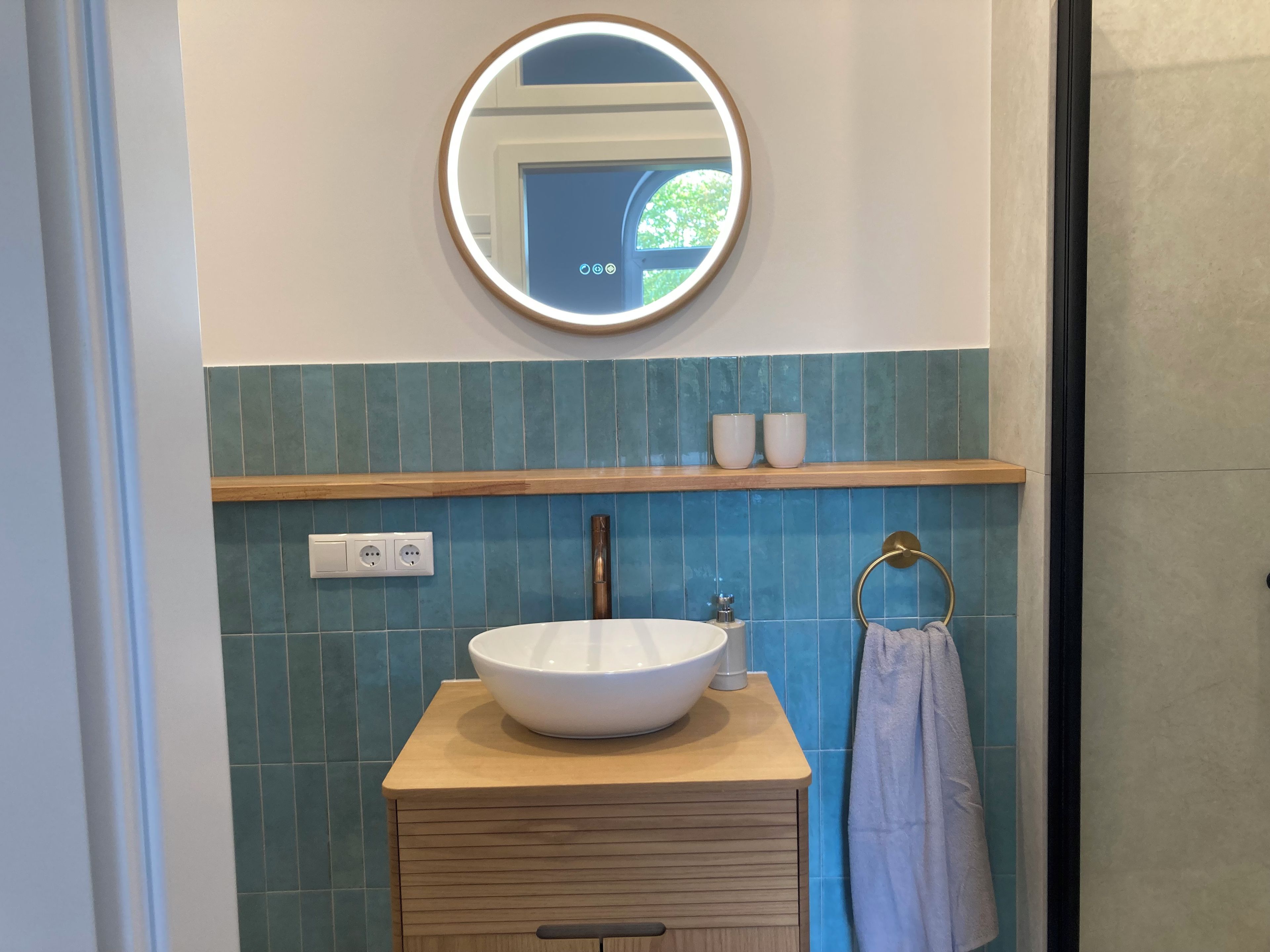 Une salle de bain avec un miroir rond, un lavabo blanc, un porte-serviettes et une étagère avec deux tasses.
