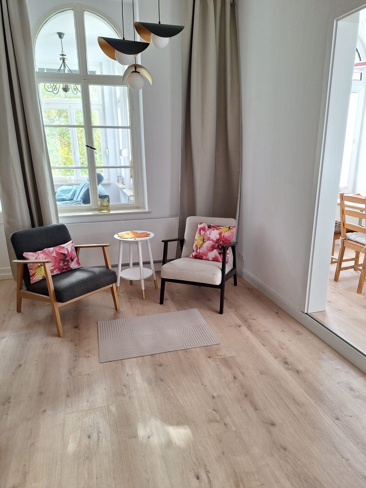 A room with a wooden floor, two chairs with floral cushions, a small white table, and a large window with curtains.