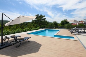 Ein rechteckiger Pool mit Fliesenbordstein, umgeben von einer Holzterrasse. Liegestühle und Sonnenschirme spenden Schatten.