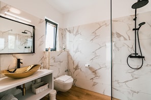 Ein modernes Badezimmer mit Marmorfliesen, einem goldenen Waschtischbecken, einer Toilette und einer Dusche mit einer Handbrause.