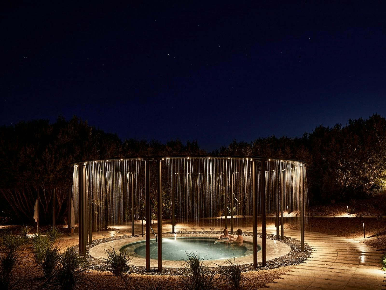 A circular outdoor pool with a central water feature, surrounded by a metal structure with vertical elements, set against a night sky.