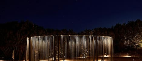 A circular outdoor pool with a central water feature, surrounded by a metal structure with vertical elements, set against a night sky.