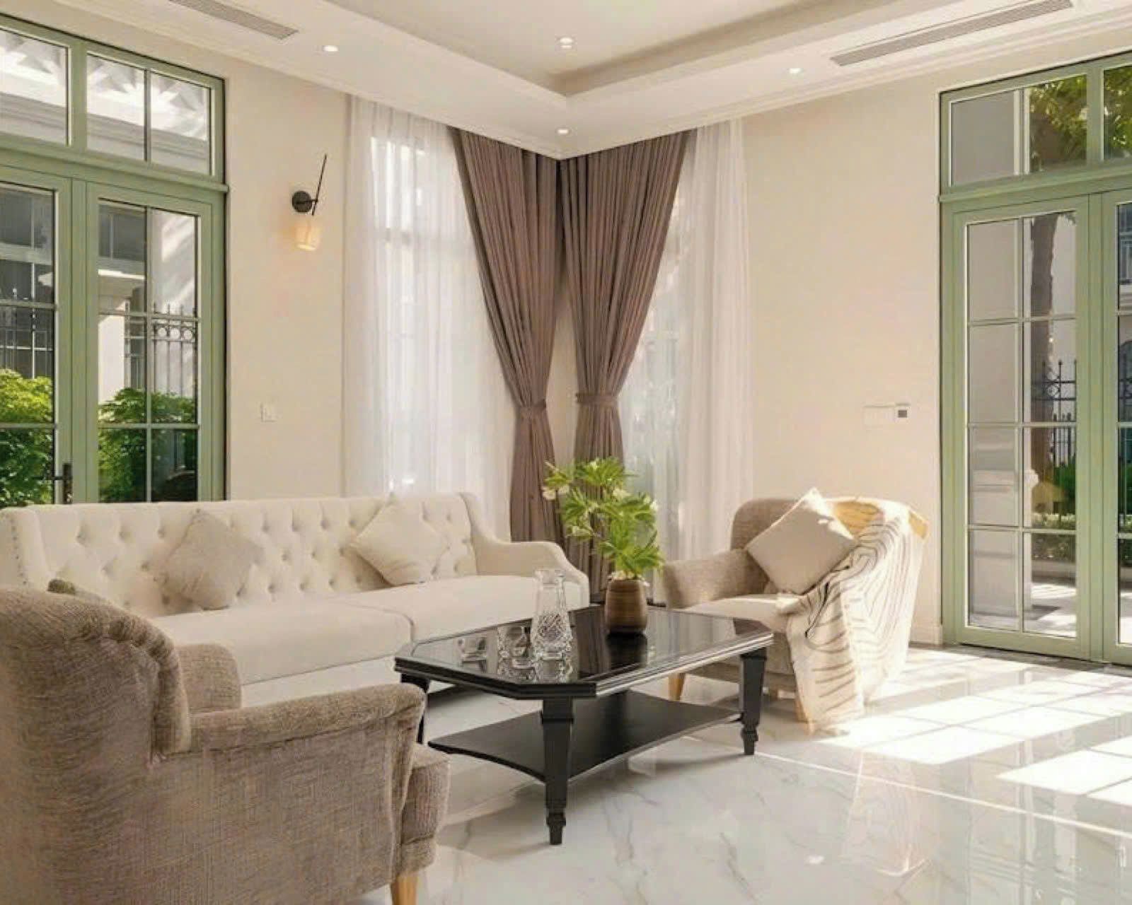 A spacious living room with a large sofa, a coffee table, and a chair. There are large windows with curtains and a view of greenery outside.