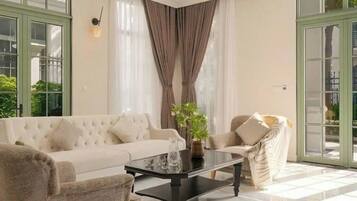 A spacious living room with a large sofa, a coffee table, and a chair. There are large windows with curtains and a view of greenery outside.