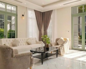 A spacious living room with a large sofa, a coffee table, and a chair. There are large windows with curtains and a view of greenery outside. - Blue Haven Tropical Villas Phu Quoc (Phu Quoc)