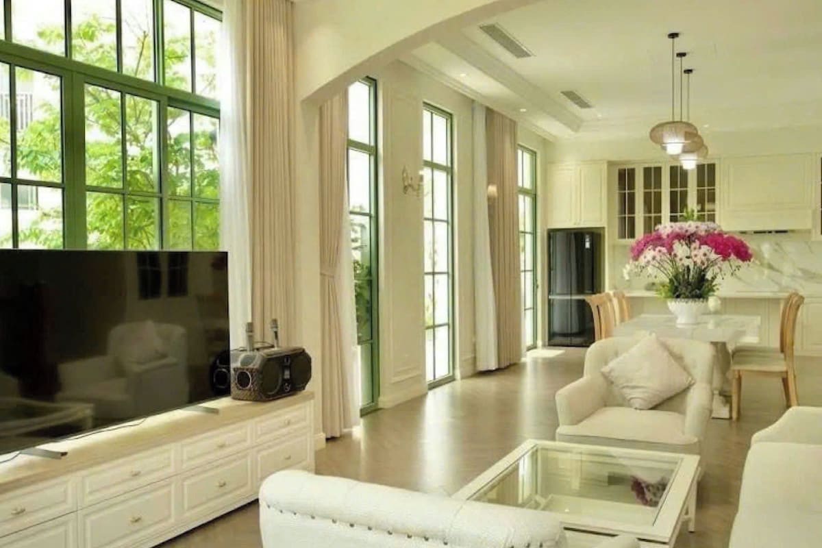 A spacious living area with a large flat-screen TV, a white sofa, a glass coffee table, and a dining area with a white table and chairs.