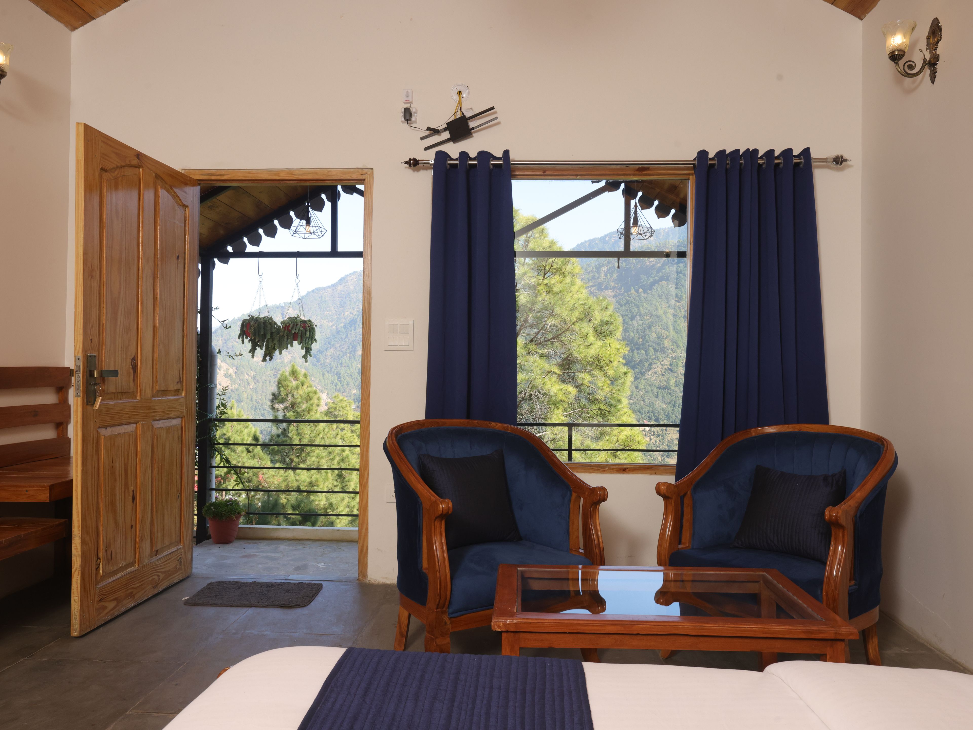 Ein Zimmer mit einem Bett, zwei Stühlen, einem Tisch, einem Fenster mit Vorhängen, einer Holztür und Blick auf Berge.