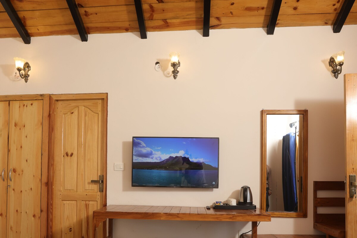 A room with a bed, a desk with a computer, a chair, a wooden wardrobe, and a wall-mounted TV.