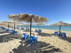 A beach with blue plastic chairs and straw umbrellas, a sandy beach, and a clear blue sky. - Cecil Hotel - Ras Sedr (Ras Sudr)