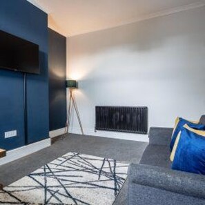 A modern living room with a dark blue accent wall, a grey sofa with blue cushions, a patterned rug, and a floor lamp. - Darlington Town Centre - Stay 7 - Near Attractions (Darlington)