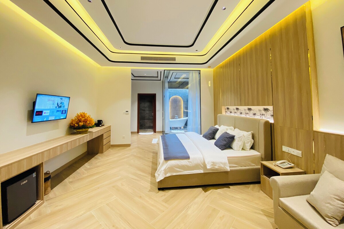 Un dormitorio moderno con cama, mesita de noche, televisor y vistas al baño.