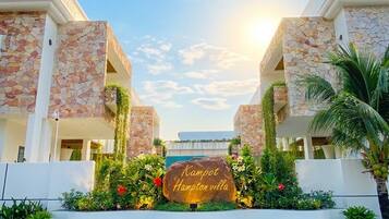 Edificio de fachada de piedra con un cartel que dice 'Kampot Hampton villa', rodeado de follaje y un cielo despejado.