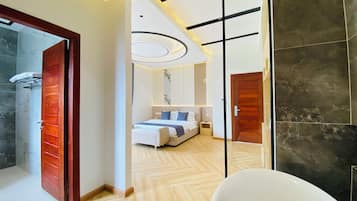 A modern hotel room with a wooden floor, a large bed with a headboard, a bedside table, a nightstand, and a bathroom with a shower area.