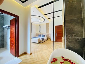 Una habitación de hotel moderna con suelo de madera, una cama grande con cabecera, una mesita de noche y un baño con zona de ducha.