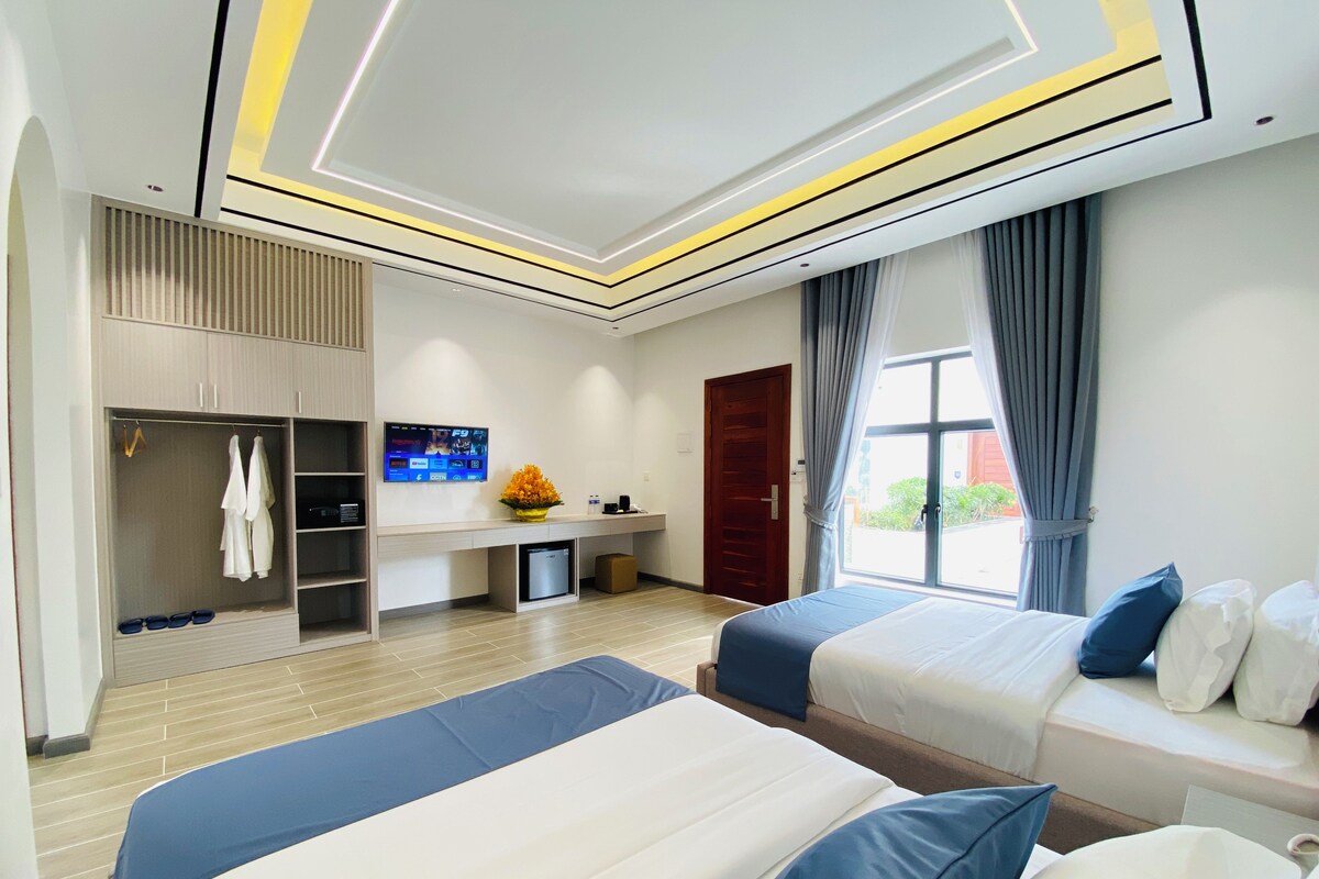 A hotel room with two beds, a desk with a TV, a wardrobe, and a window with curtains.