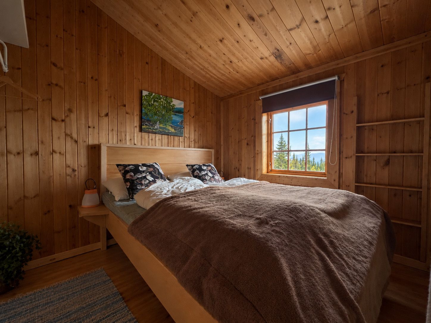 Ein Schlafzimmer mit Holzwänden, einem Bett, einem Nachttisch, einem Fenster mit Blick und einem Bild an der Wand.