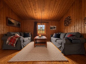 Una habitación acogedora de estilo cabaña con dos sofás grises, techo de madera, alfombra y ventana con vista a árboles.
